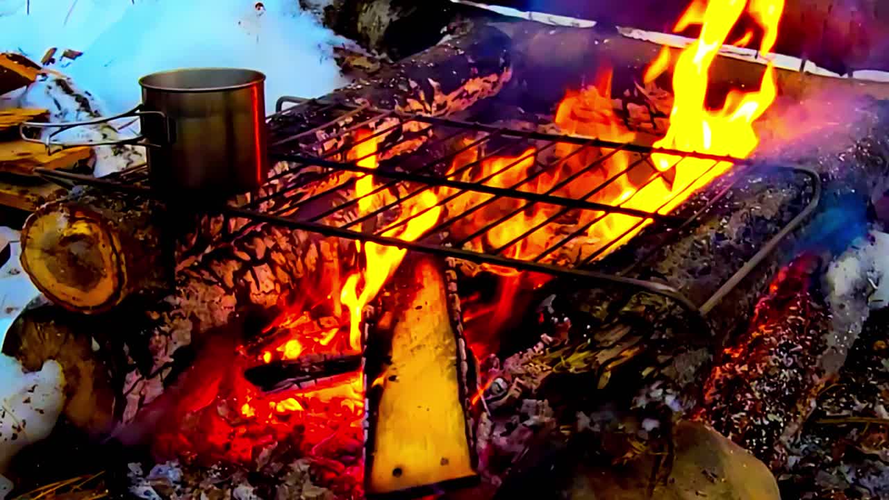 hoguera estilo cabaña de troncos hecha a mano con una parrilla de metal sobre ella hirviendo café con chocolate caliente humeando con fuego viger ardiendo con tablones de madera contrachapada alimentados en el fuego en un terreno cubierto de nieve invernal