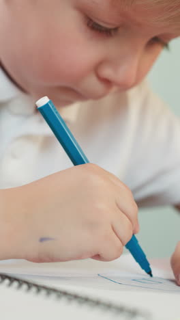 little boy puts marker cap and draws picture on copybook page sitting at table closeup. talented focused toddler pupil does tasks at art lesson sticking out tongue