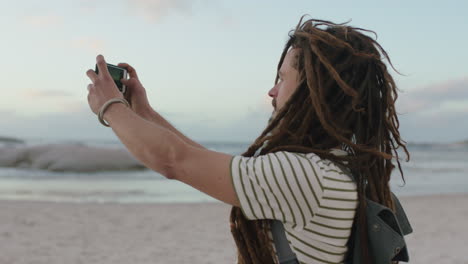 Junger,-Unbeschwerter-Mann-Mit-Dreadlocks,-Der-Am-Strand-Mit-Dem-Telefon-Fotos-Macht-Und-Ein-Gestreiftes-Hemd-Trägt