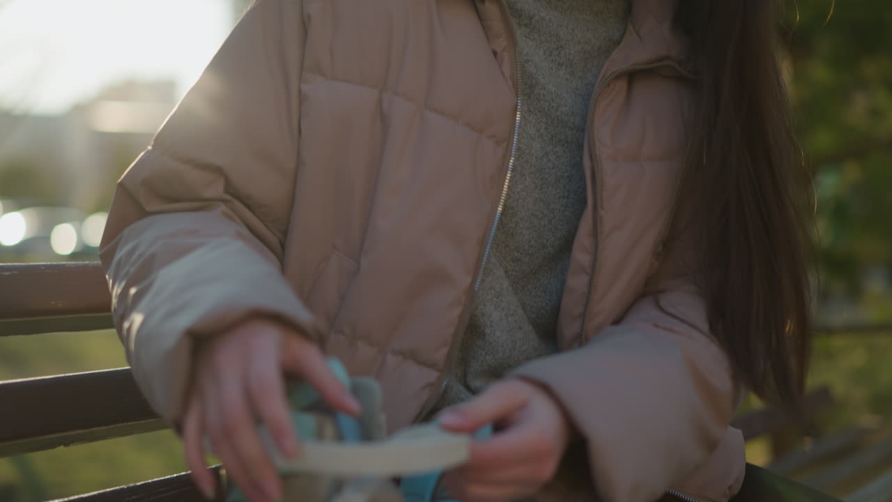 молодая женщина в персиковой куртке сидит на скамейке в открытом парке, снимая свои ролики. кадр сосредоточен на ее верхней части тела, показывая детали ее одежды и действия