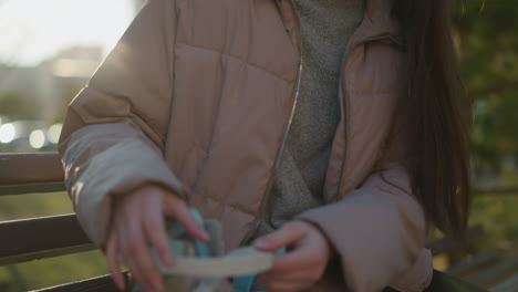 una mujer joven con una chaqueta de melocotón está sentada en un banco en un parque al aire libre, quitando sus patines. la toma se centra en su parte superior del cuerpo, mostrando los detalles de su ropa y la acción