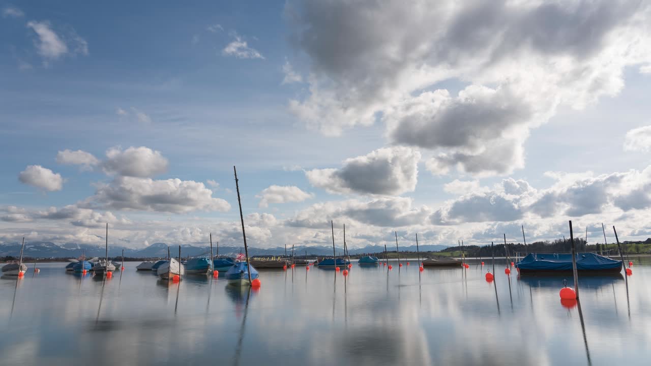 Spring is coming. Clouds are drifting across the blue sky, and the sun is shining. Fishing boats are bobbing on the calm lake. The mountain ranges are in the background. clouds reflected in the water.