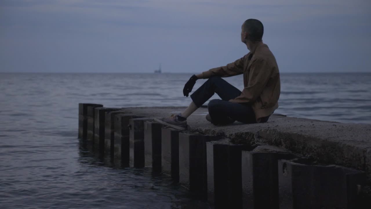 Young Man Relaxing While Sitting Alone On A Wharf At The Beach - Medium Shot