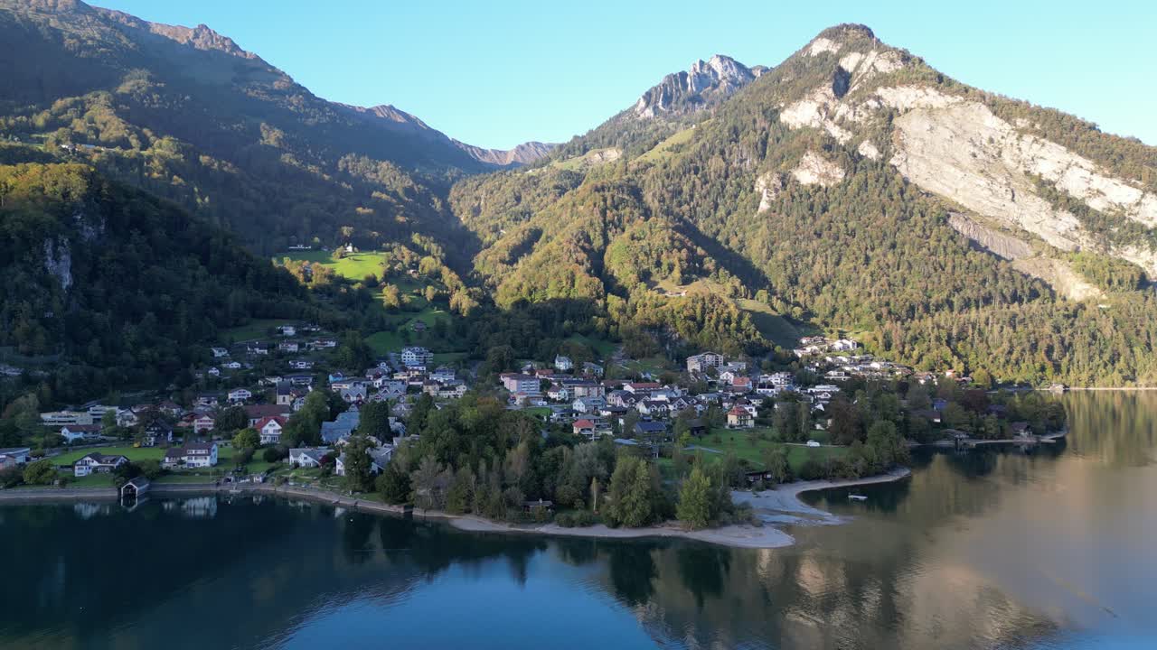 Flyover of a small marina and residential houses above Walensee, with the majestic green-dressed mountain Alps beautifully reflected in the water on a clear day