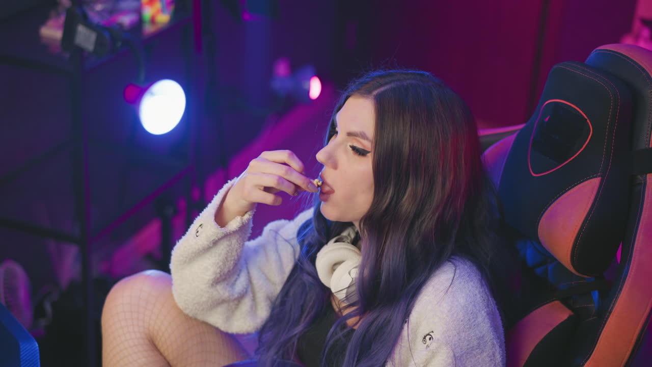 Gamer seated in comfortable gaming chair with legs crossed, enjoying popcorn under soft purple and pink lighting, immersed in relaxation and leisure mood inside tech-inspired studio setup