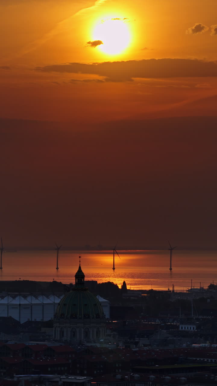 Aerial drone view over Copenhagen, with the Marble Church dome in the foreground and offshore wind turbines glowing in the golden light of dawn. Vertical