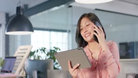 Glückliche-Asiatische-Geschäftsfrau,-Die-Ein-Tablet-Benutzt,-Auf-Dem-Smartphone-Spricht-Und-Im-Büro-Lacht,-In-Zeitlupe