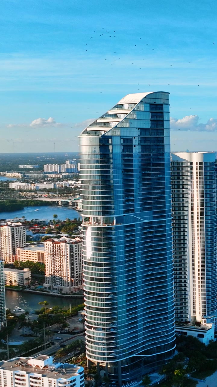 Luce del mattino su un moderno grattacielo di vetro a Sunny Isles Beach, Miami. Punto di vista aereo di una facciata architettonica curva, un uccello in volo, una spiaggia sabbiosa e l'orizzonte blu dell'oceano. Video verticale