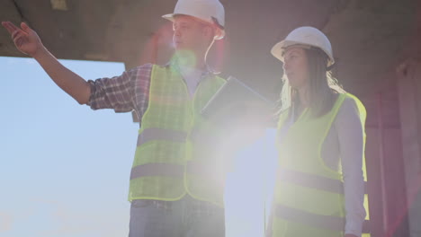 trabajador de la construcción hombre y arquitecto mujer en un casco discutir el plan de construcción de la casa hablar el uno al otro sobre el diseño sosteniendo una tableta mirar los dibujos fondo de los rayos del sol.