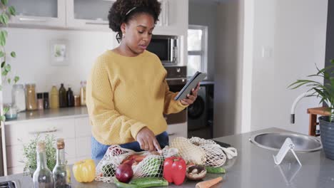 happy african american woman using tablet and unpacking groceries in kitchen, slow motion