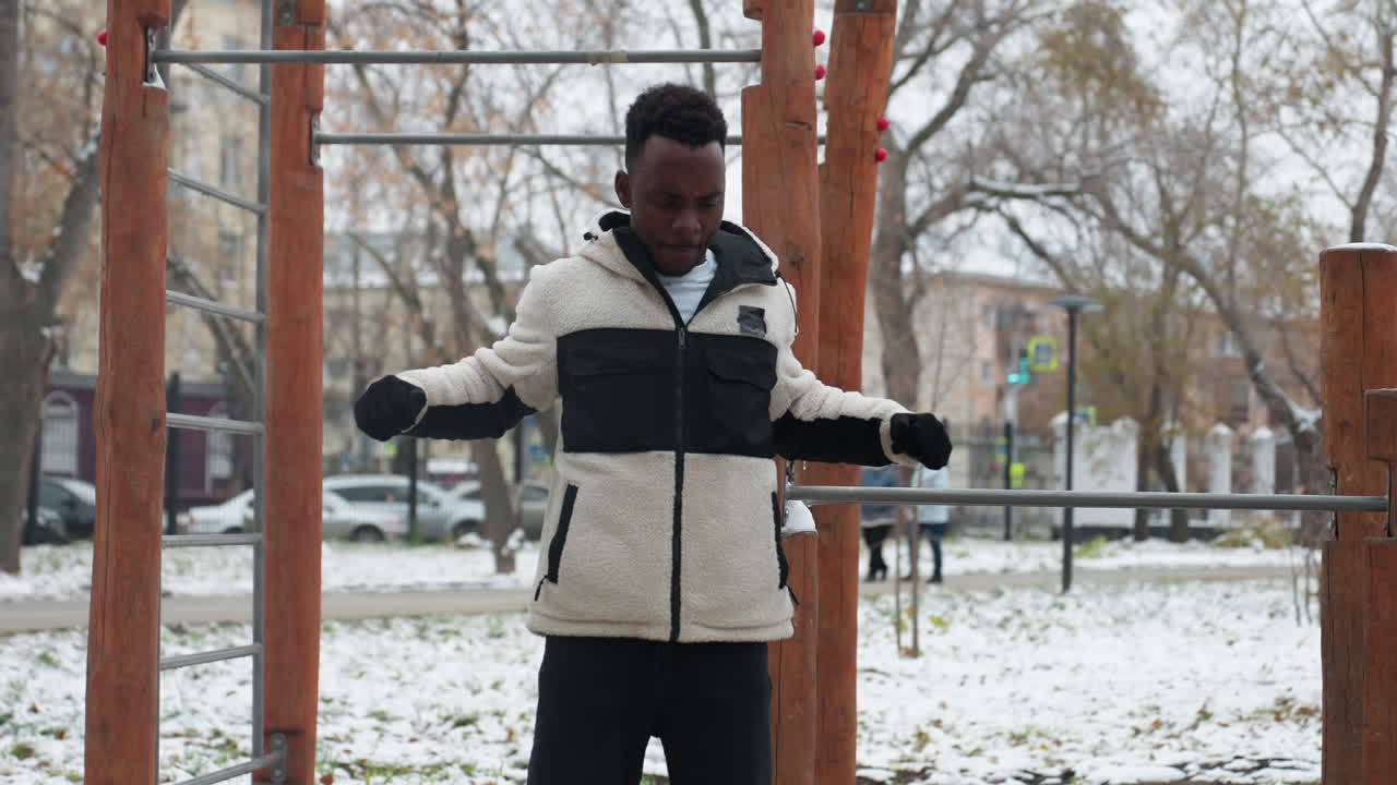 Sportsman working out doing arm stretch outdoors in winter, snow-covered ground with workout equipment and people walking blurred in the background