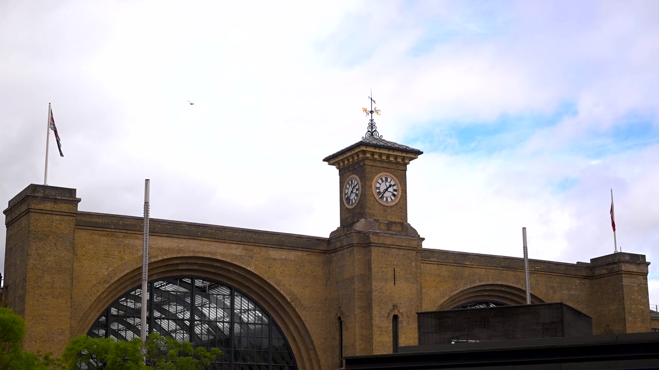 La torre dell'orologio della Stazione di King's Cross e il tetto a volta del capannone ferroviario si ergono contro un cielo nuvoloso con bandiere e un uccello di passaggio.