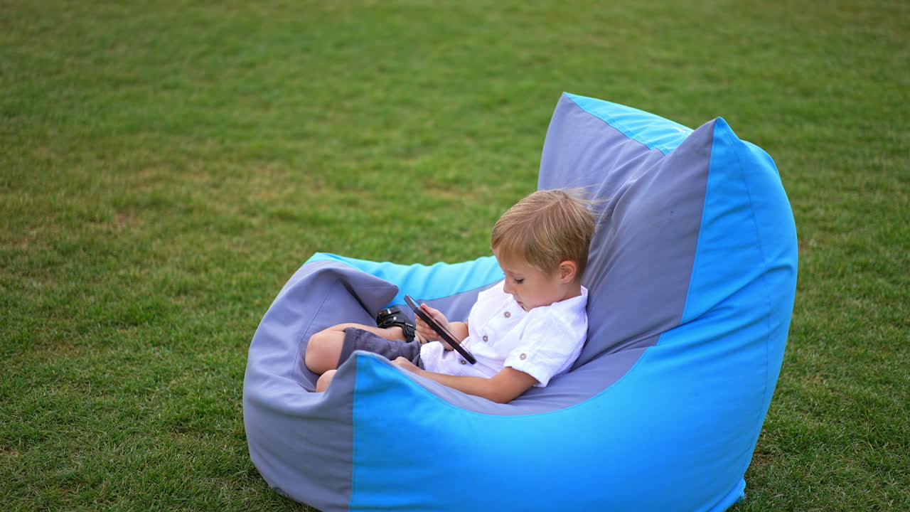 Blue-eyed blond boy of six years having rest outdoors. Lovely kid using smartphone sitting in bean bag chair on the green grass.