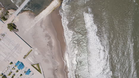 aerial-top-down-view-of-Puerto-Colombia-Dock-unfolds-with-waves-gracefully-crashing-on-the-shore-and-the-beach