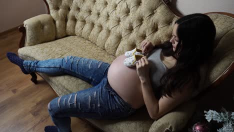 Pregnant-woman-play-with-baby-shoes-on-cozy-couch-near-Christmas-tree