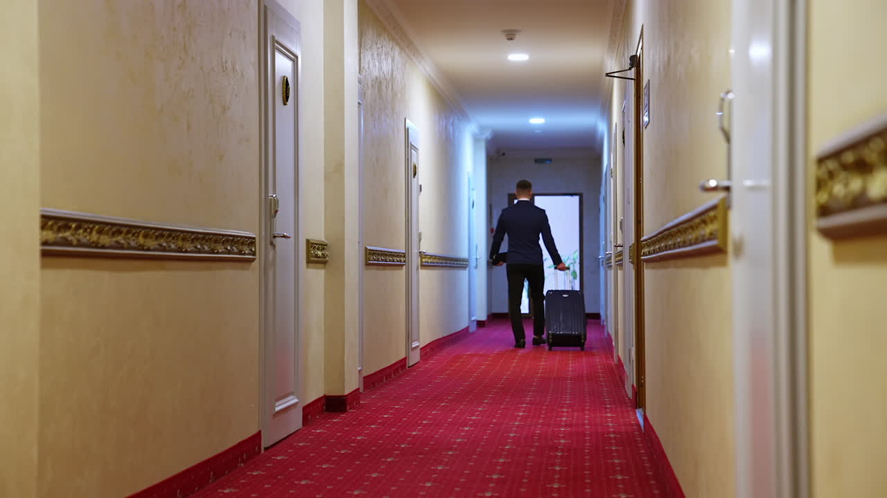 Back view of businessman walking with suitcase in hotel corridor. Modern interior with white walls, doors and red carpet.