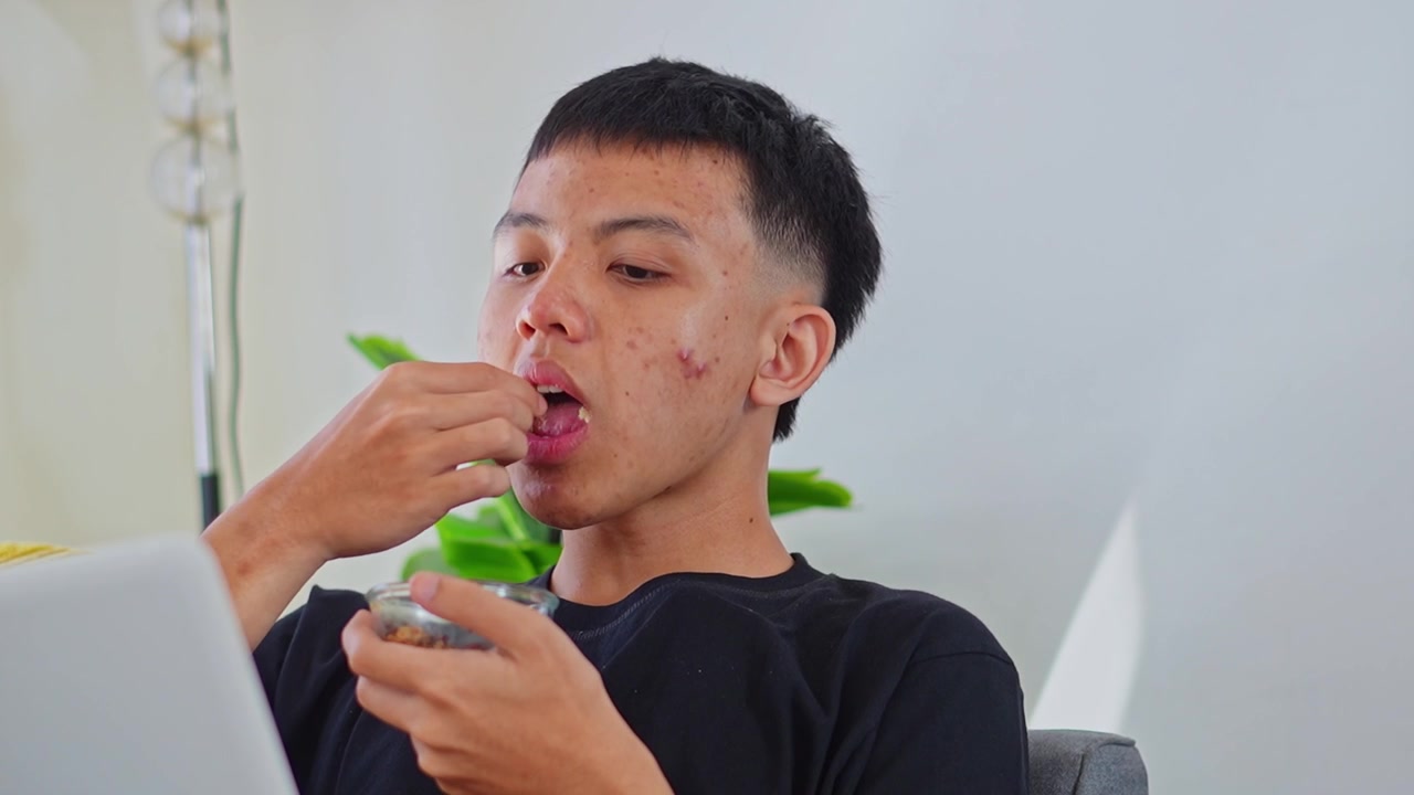 Young man using laptop while eating crunchy granola from small glass bowl at home