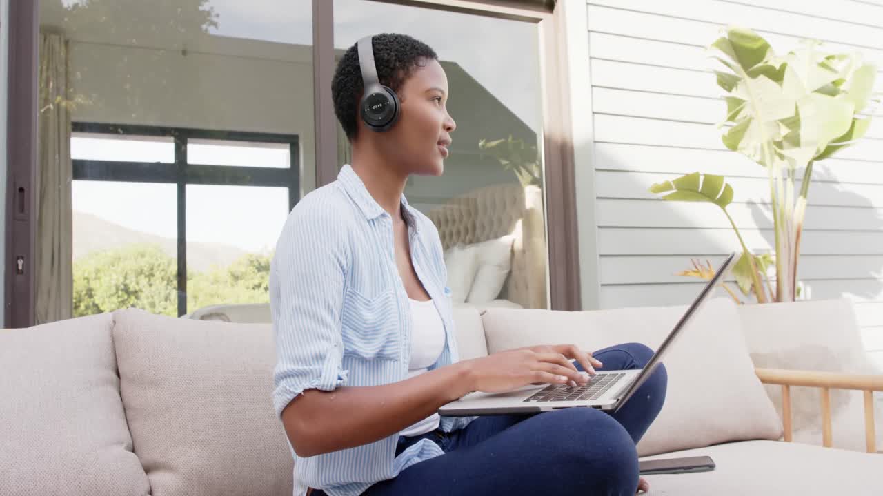 African american woman sitting on sofa on terrace wearing headphones, using laptop, slow motion