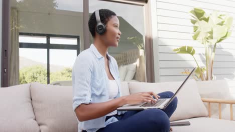 Mujer-Afroamericana-Sentada-En-Un-Sofá-En-La-Terraza-Usando-Auriculares,-Usando-Una-Computadora-Portátil,-Cámara-Lenta