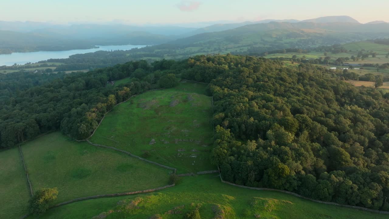 Woodland covered hilltop with camera rise revealing fielded green lakeside valley and mountains beyond at sunrise. Summer. Windermere, Cumbria, UK