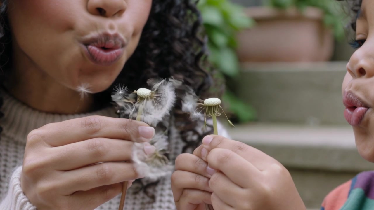 Blowing mother and child in cream-knit, striped knits on steps, pursed lips blowing dandelion puffs