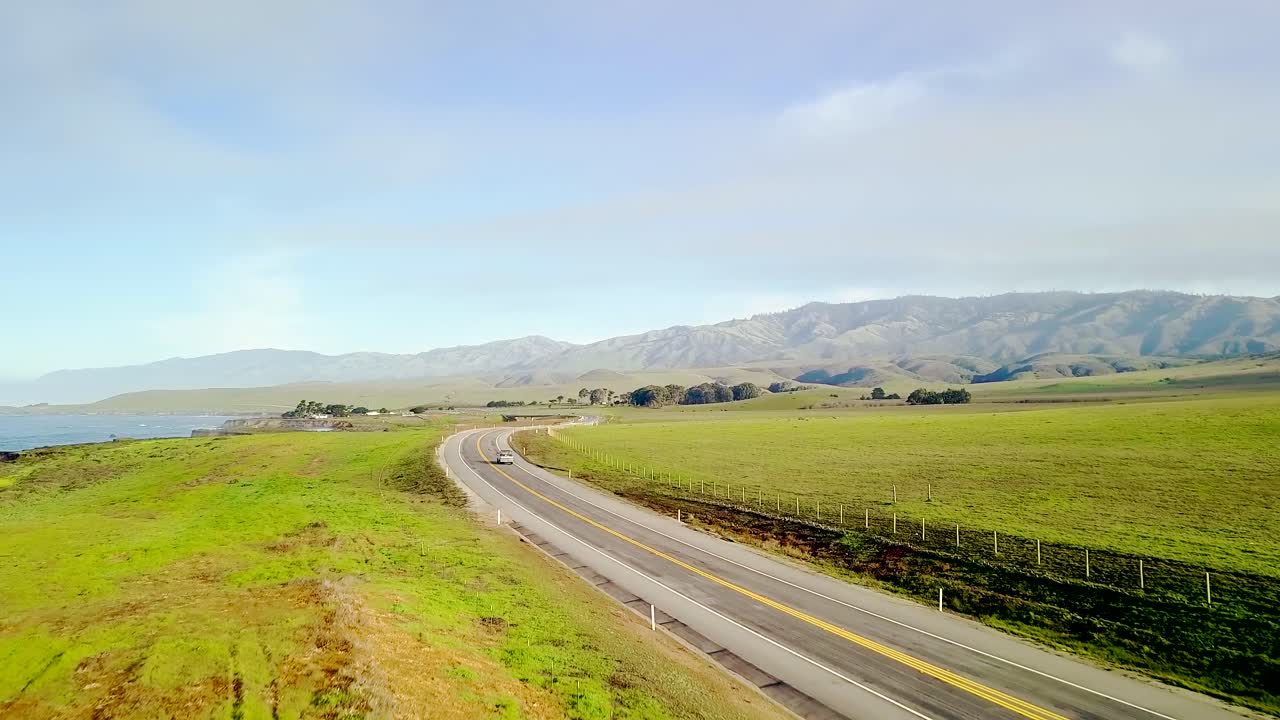 구불구불한 캘리포니아 해안 고속도로와 구불구불한 푸른 언덕의 공중 광활한 전망은 바다 해변 절벽을 드러내기 위해 상승하는 빠른 자동차와 일출 시 파도가 부서지는 파도를 따라 올라갑니다.