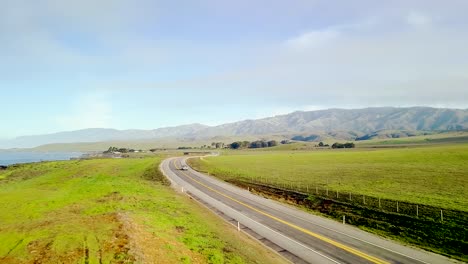 Aerial-wide-view-of-winding-California-coast-highway-and-rolling-green-hills-following-fast-cars-rising-to-reveal-ocean-beach-cliffs-and-crashing-waves-surf-at-sunrise