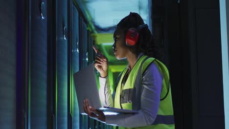 técnica informática afroamericana que usa una computadora portátil trabajando en la sala de servidores de negocios