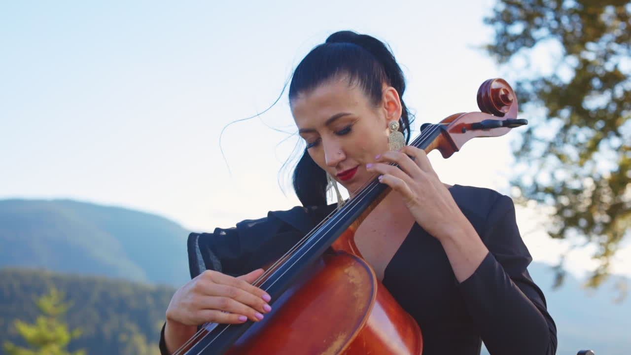 Dark-haired female musician uses no bow to play violoncello. Woman moving her head in the rhythm of music. Mountains backdrop in blur.