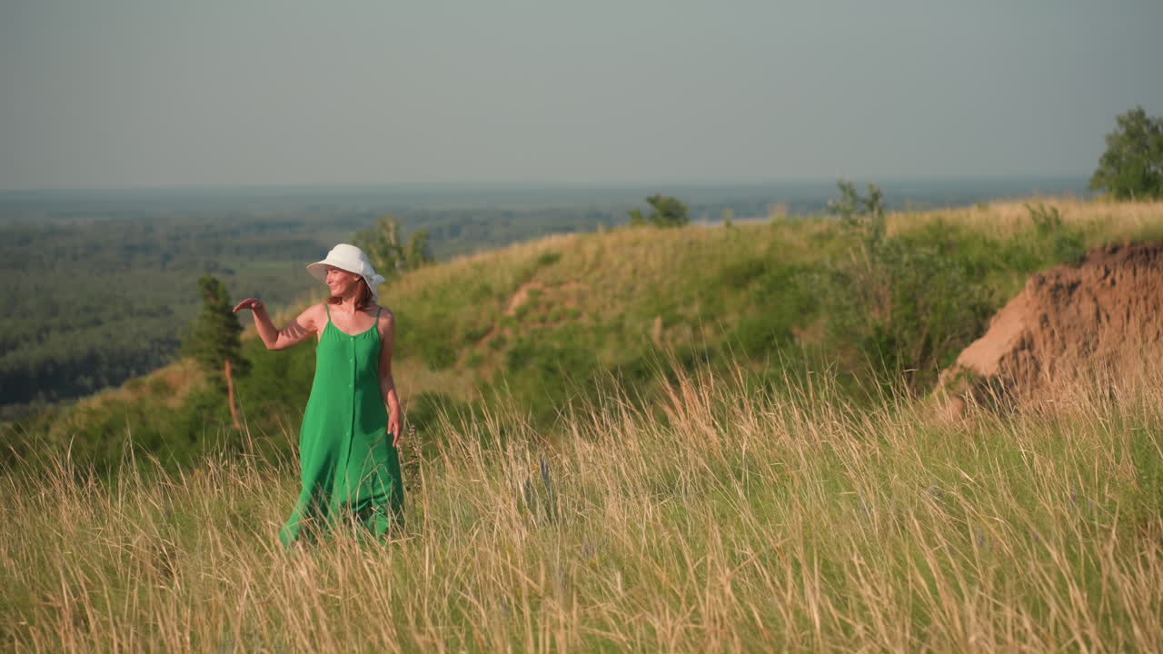 woman in green gown and white sunhat turns to look back while walking along grassy cliffside under warm light, with lush hills, river in distance, and wind gently swaying dry
