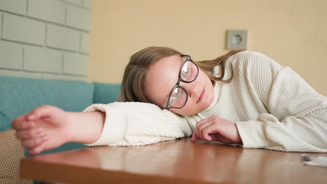 young woman wearing glasses and white sweater rests head on wooden table with soft smile while gently drawing or tracing shapes with finger
