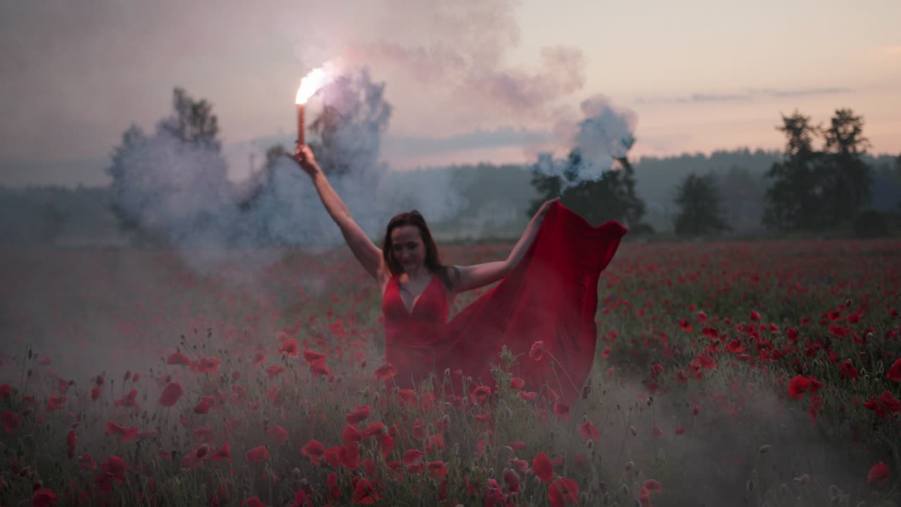 Ritratto di una graziosa ragazza bruna in abito rosso che balla con un fumogeno rosso acceso in un campo di papaveri.