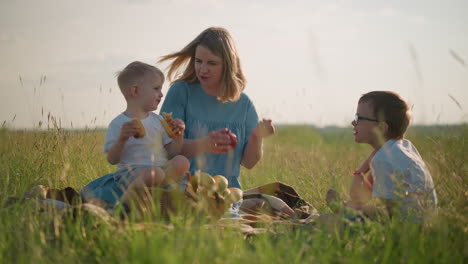 a mother in a blue dress sits on the grass with her two sons, both wearing white tops, enjoying snacks together. the mother holds a red apple while one child reaches out