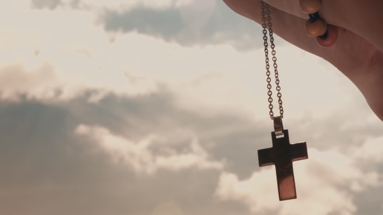 Mano che tiene una collana con croce cristiana contro un cielo nuvoloso