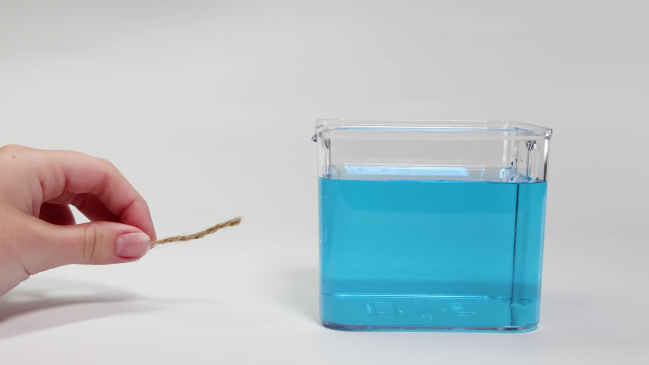A hand places a string in a water-filled container, observing its buoyancy. Bright lighting highlights the clear, blue liquid