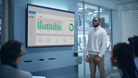 Office Conference Room Meeting Presentation: Black Businessman Talks, Uses Wall TV to Show Company Growth with Big Data Analysis, Graphs, Charts, Infographics. Multi-Ethnic e-Commerce Startup Workers