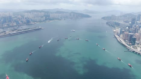 Convoy-of-local-Fishing-boats-causing-in-Hong-Kong-Victoria-bay,-with-city-skyline-in-the-horizon,-Aerial-view