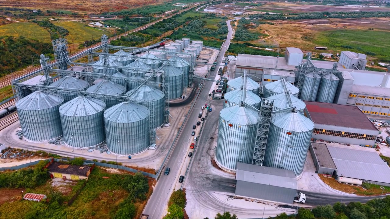 Industrial grain terminal with multiple storage silos. Elevated drone view of metal bins, conveyor towers, factory buildings, rail tracks, roads and parked vehicles.