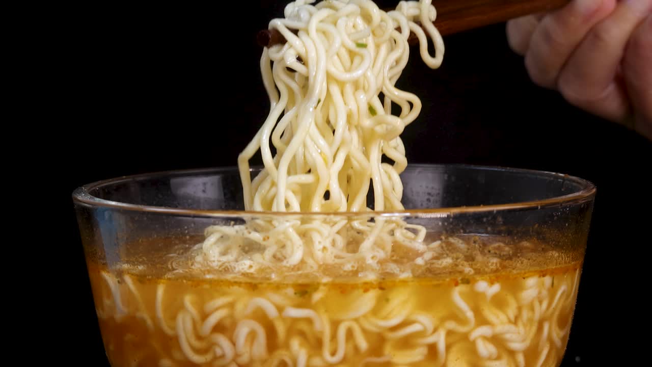 Hand lifts steaming instant noodles with chopsticks from glass bowl of broth, dramatic black background