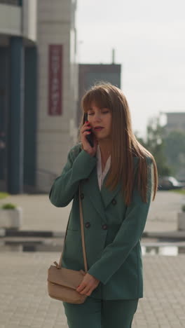 disgruntled adult woman with long red hair argues on mobile phone walking on sidewalk in city on cloudy day. busy mother scolds child. family dispute