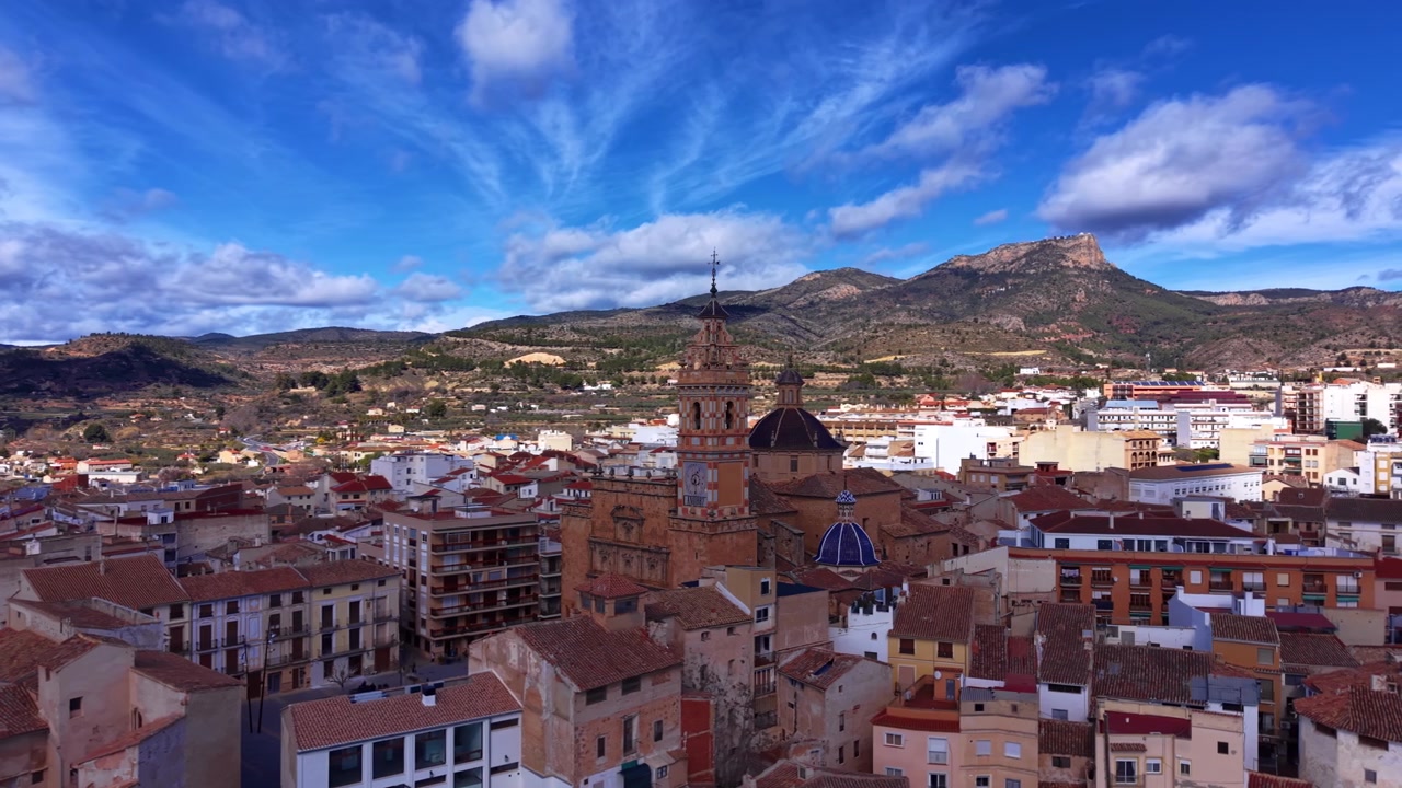 Vista panoramica di Chelva, Spagna, con una torre di chiesa barocca e una cupola blu che si ergono sopra i tetti in terracotta, con montagne e cielo blu sullo sfondo