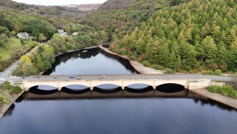 Experience the tranquil beauty of Ladybower Reservoir in the Peak District, England. A scenic bridge spans the vast water, surrounded by lush forested hills, offering a serene glimpse into this area.