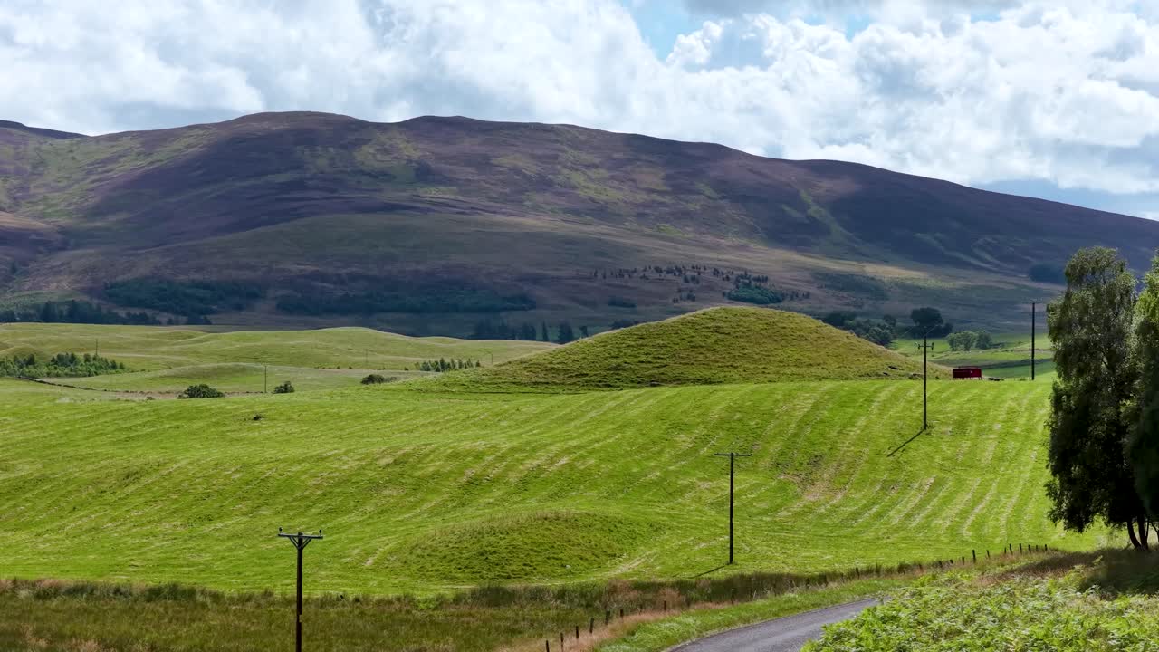 Camera glides down winding countryside road, revealing green meadows, rolling hills, and distant mountains
