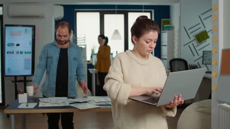 Portrait-of-casual-woman-using-laptop-and-smiling-at-camera-while-coworker-holding-cup-looks-at-papers