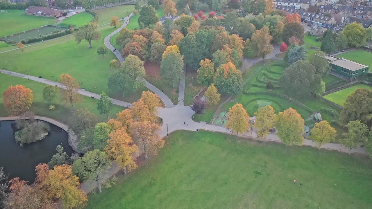 Static aerial shot highlights geometric paths and vibrant fall foliage of Hillsborough Park, with a sunlit green lawn, pond, and winding trails inviting visitors to relax on Sheffield’s northwest side