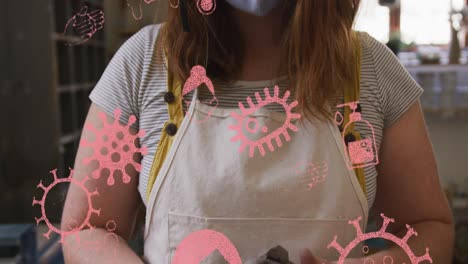 Animation-of-covid-icons-and-cells-over-woman-wearing-face-mask-holding-clay-in-pottery-workshop