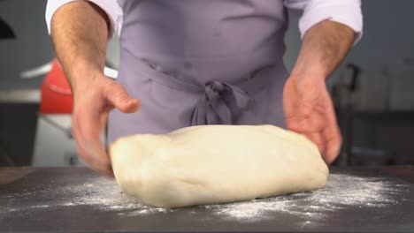 a baker at work, wearing a gray apron, kneading a big dough with his hands in order to prepare bread, focaccia or pizza