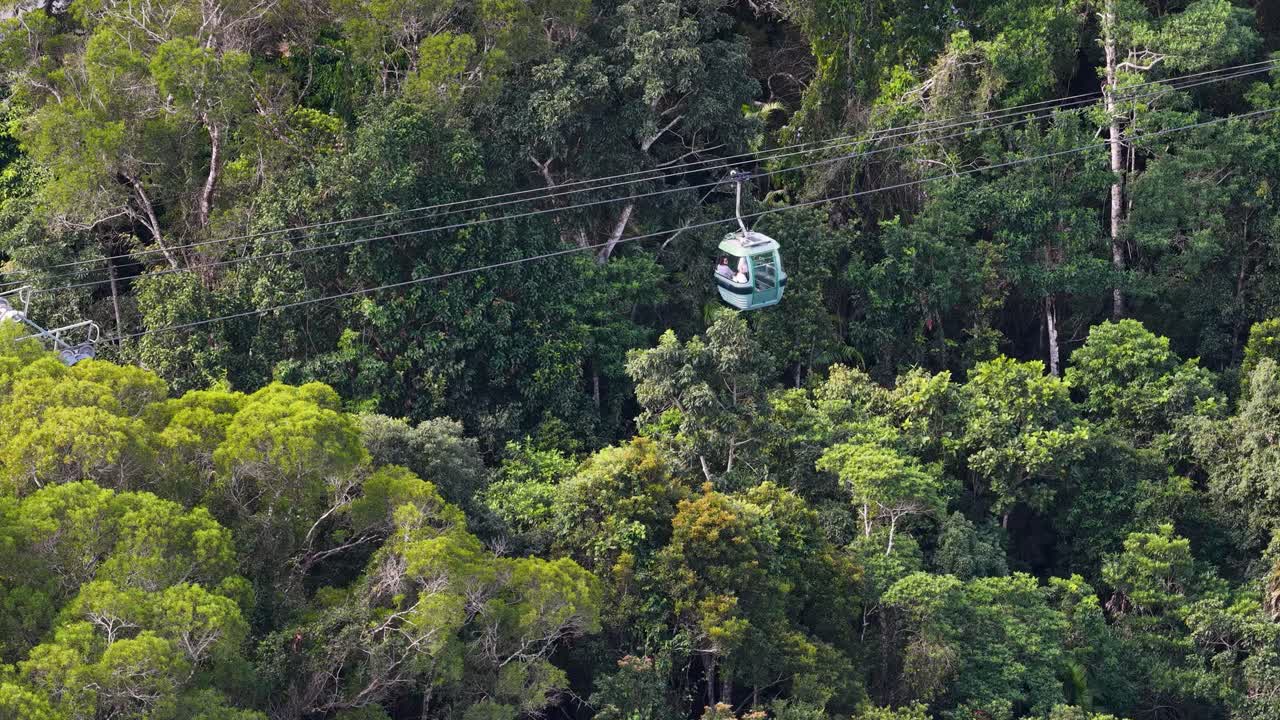 Aerial view of a cable car traveling smoothly over dense, sunlit rainforest. The camera tracks the gondola’s movement, highlighting vibrant green foliage and natural landscape