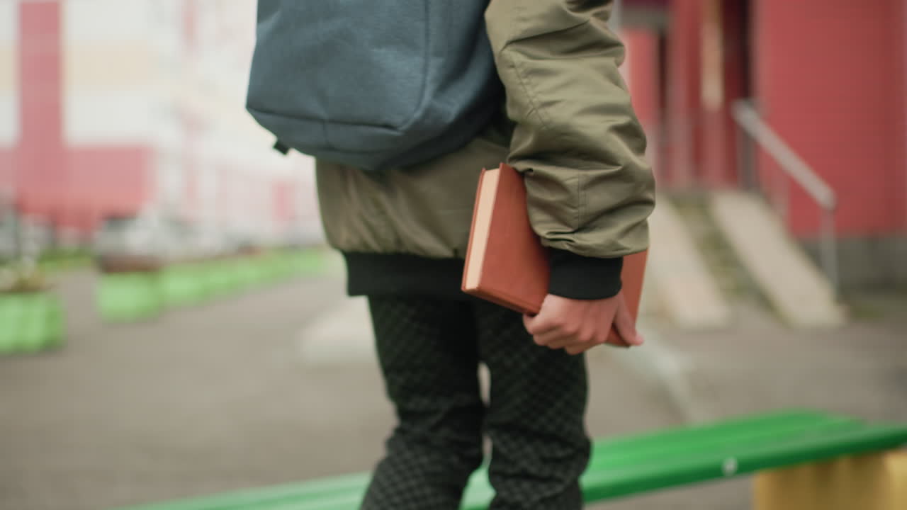 Close up rear view of kid carrying backpack and holding book while walking toward outdoor bench sit and read with blurred urban background of buildings cars and colorful street decorations