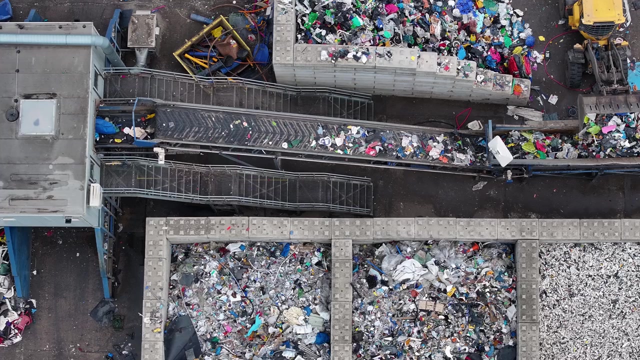 Plastic waste enters the facility for sorting and processing. Large piles of recyclable materials are visible outside. Workers process waste for resource recovery in an industrial environment.